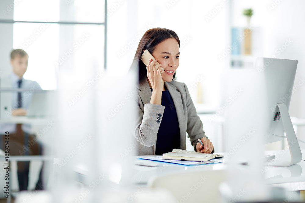 Portrait of successful Asian businesswoman busy working at desk in modern office, making calls using smartphone and smiling looking at computer screen Portrait of successful Asian businesswoman busy working at desk in modern office, making calls using smartphone and smiling looking at computer screen