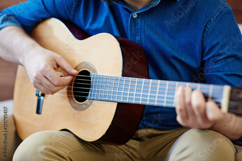 Close-up shot of man sitting on comfortable armchair and practicing in playing acoustic guitar