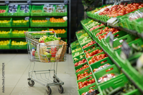 Metal shopping trolley with healthy grocery items standing in fruit and vegetable department of hypermarket