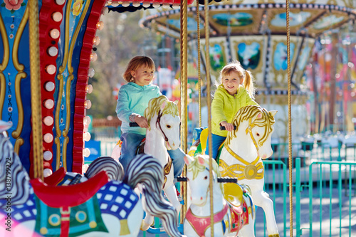 Cute little sisters enjoying spring in funfair: they riding on colorful carousel and looking at camera with wide smiles