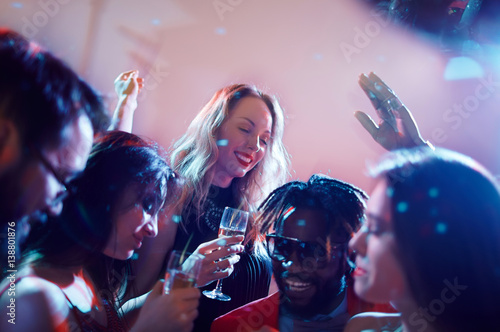 Joyful multiethnic friends holding champagne flutes in hands and dancing with raised hands in trendy night club