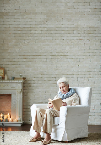Serious elderly woman wrapped up in reading adventure novel while sitting by fireplace decorated with burning candles