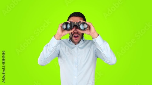 Astonished young man using a binoculars against green background