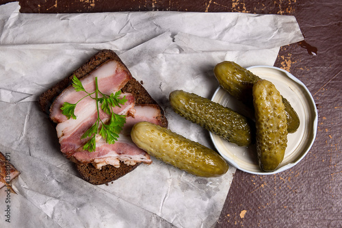 Slices of smoked bacon with rye black bread and several pickles on the white packaging paper.