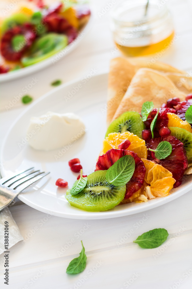 Thin crepes with fresh kiwi, orange, sicilian orange, honey, cream and mint leaves on white rustic wooden table. Selective focus Thin crepes with fresh kiwi, orange, sicilian orange, honey, cream and mint leaves on white rustic wooden table. Selective focus