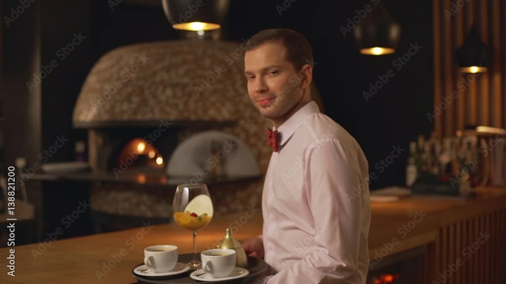 Middle-aged Caucasian male waiter in white shirt and bow tietaking tray with cups and glass smiling showing ok in slowmotion Middle-aged Caucasian male waiter in white shirt and bow tietaking tray with cups and glass smiling showing ok in slowmotion
