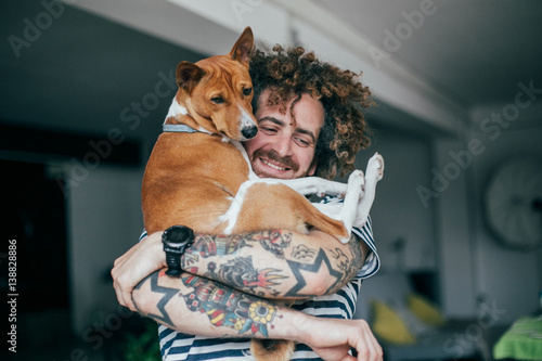 Crazy haired hipster in striped shirt and tattoos hugging dog in industrial loft apartment