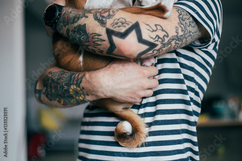 Hipster with tattoos holding purebred dog in loving arms with striped shirt.