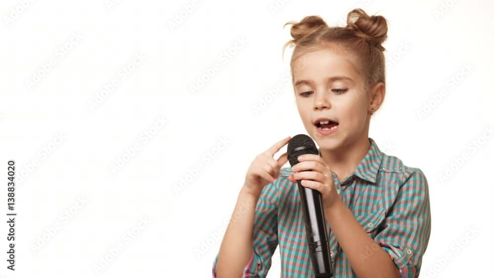 Charming Caucasian small kid girl in pladi checking black microphone then singing on it smiling on white background Charming Caucasian small kid girl in pladi checking black microphone then singing on it smiling on white background
