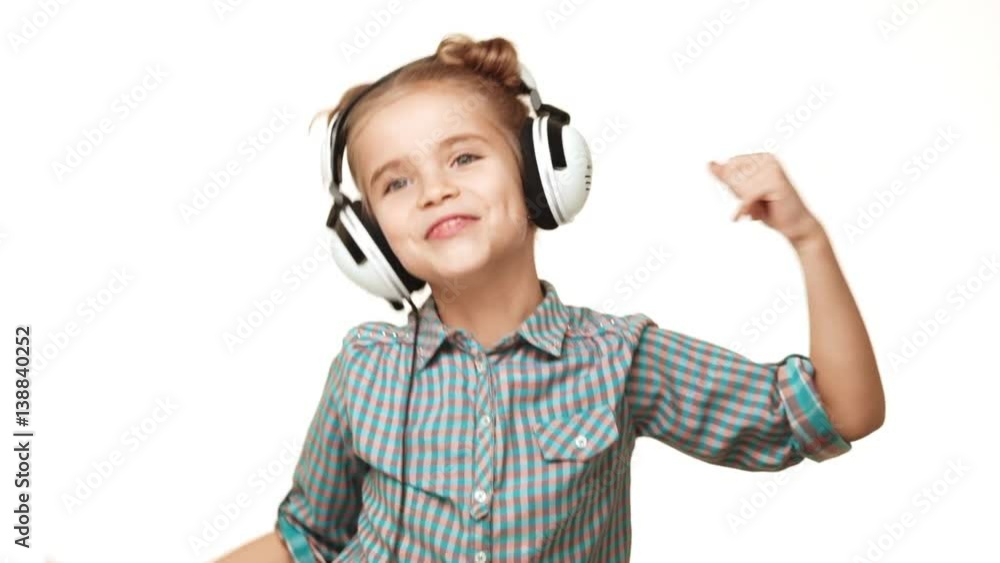 Small active Caucasian kid girl in plaid shirt and hair buns listening music through headphones and dancing on white background in slowmotion Small active Caucasian kid girl in plaid shirt and hair buns listening music through headphones and dancing on white background in slowmotion
