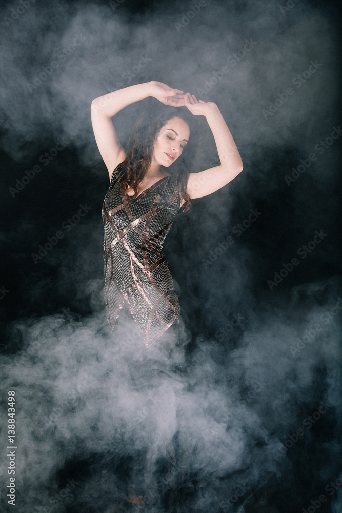 Beautiful brunette woman in a dress dancing and posing in Studio on dark background with smoke