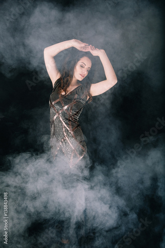Beautiful brunette woman in a dress dancing and posing in Studio on dark background with smoke