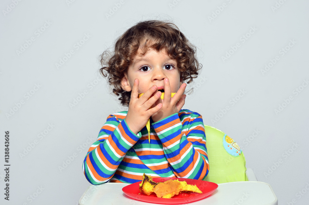 Kid getting messy while eating a chocolate cake. Beautiful curly hair boy eating sweets. Toddler in high chair being hungry.