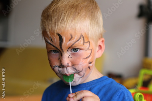 Boy with orange lion painted on his face licking lollipop