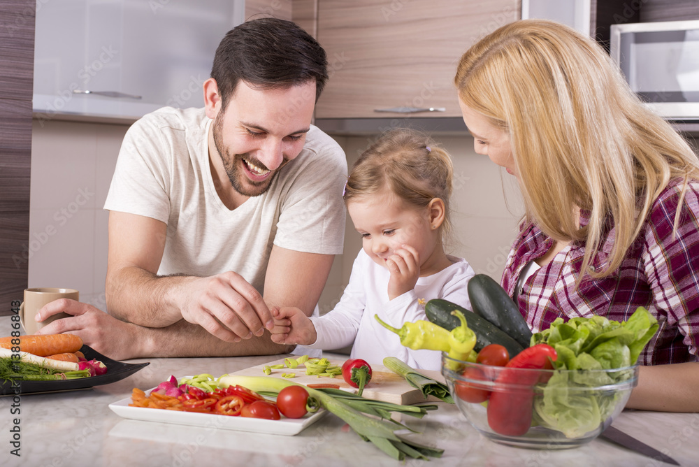 Young couple with kid having fun in kitchen while preparing fresh vegetable salad