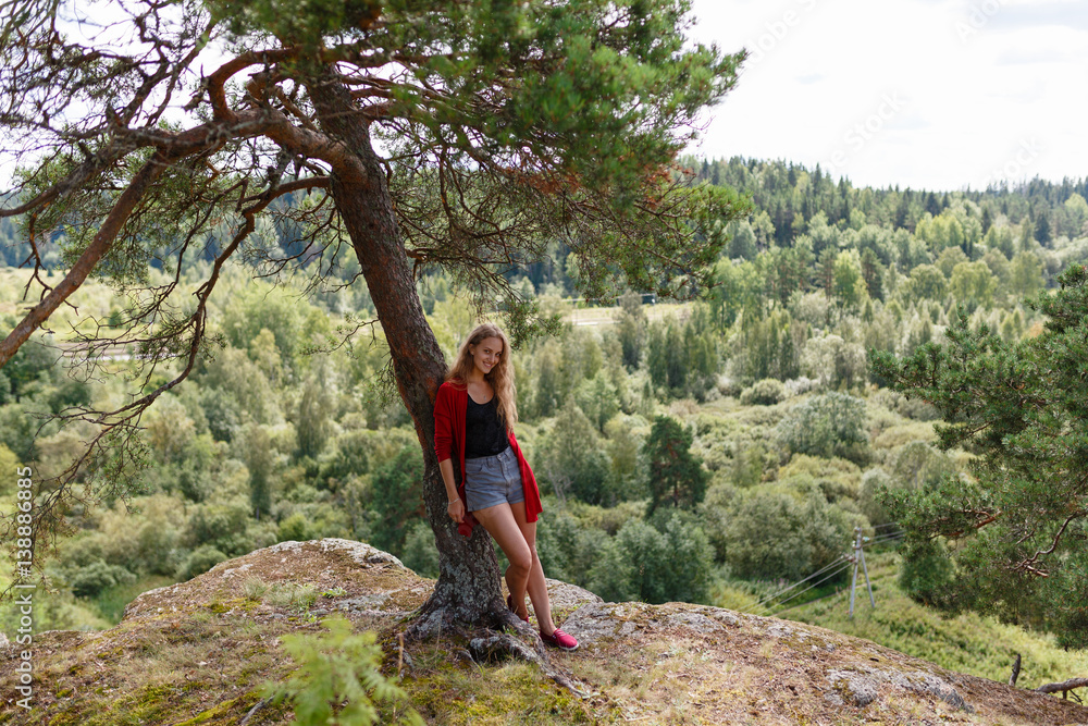 The girl in the red sweater and shorts standing at the cliff leaning on a tree and enjoy the landscape and feel the freedom. Looking at the camera and smiling.