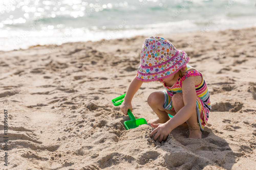 Little girl in a hat and sundress plays with a toy shovel on the beach Little girl in a hat and sundress plays with a toy shovel on the beach