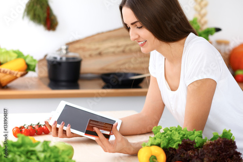 Beautiful Hispanic woman is making online shopping by tablet computer and credit card. Housewife found new recipe for cooking in a kitchen