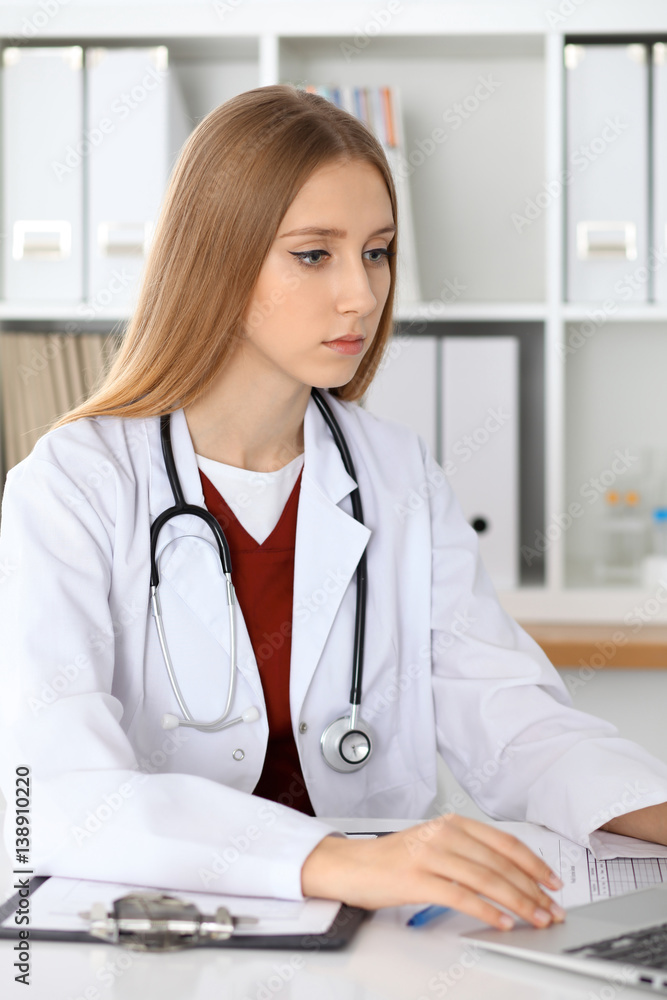 Doctor woman filling up medical form or typing on laptop while sitting at the table near the window. Medicine and health care concept Doctor woman filling up medical form or typing on laptop while sitting at the table near the window. Medicine and health care concept