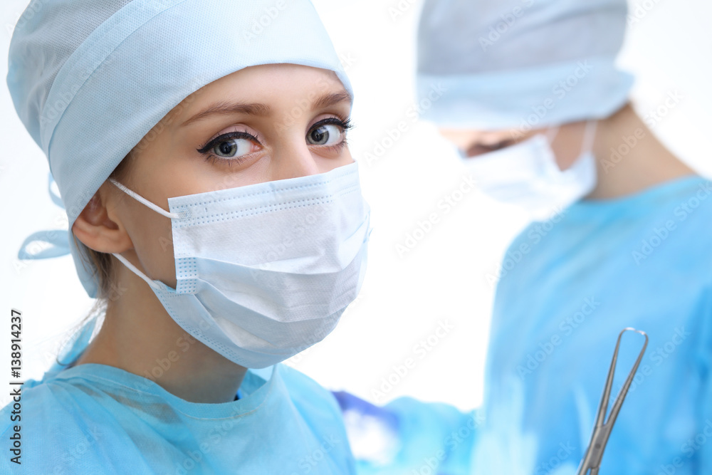 Close up of surgeon woman looking at camera while colleagues performing in background in operation room, isolated. Surgery and medicine concept Close up of surgeon woman looking at camera while colleagues performing in background in operation room, isolated. Surgery and medicine concept