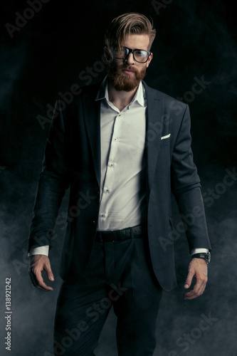 Stylish handsome bearded man in trendy suit and eyeglasses posing in smoke