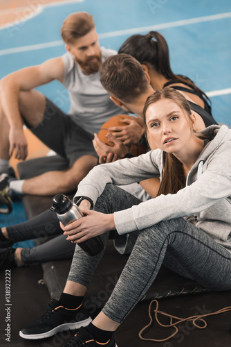 Young sporty woman holding bottle while sitting near teammates and looking at camera