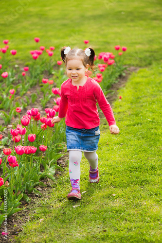 Cute funny active girl of four years old running alone cheerfully outside along flowerbed with pink tulips in spring sunny city park. Vertical color photography.