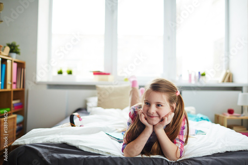 Pretty long-haired girl with ponytails lying on bed with head leaned in her hands, looking to the side and smiling mischievously