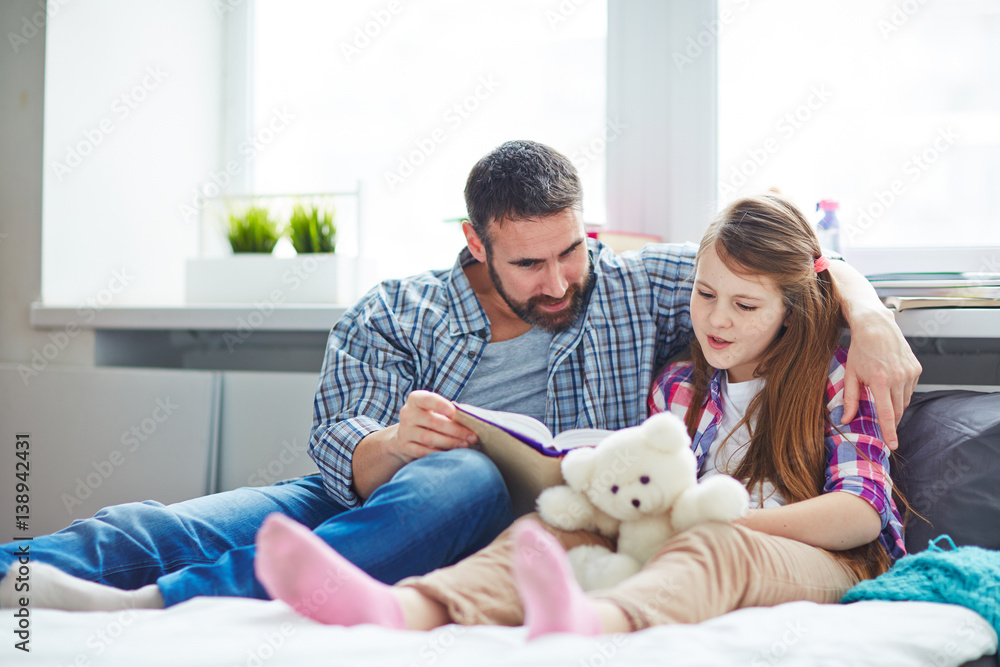 Full-length portrait of pretty family: little girl holding teddy bear in hands while reading adventure story to her bearded middle-aged father Full-length portrait of pretty family: little girl holding teddy bear in hands while reading adventure story to her bearded middle-aged father