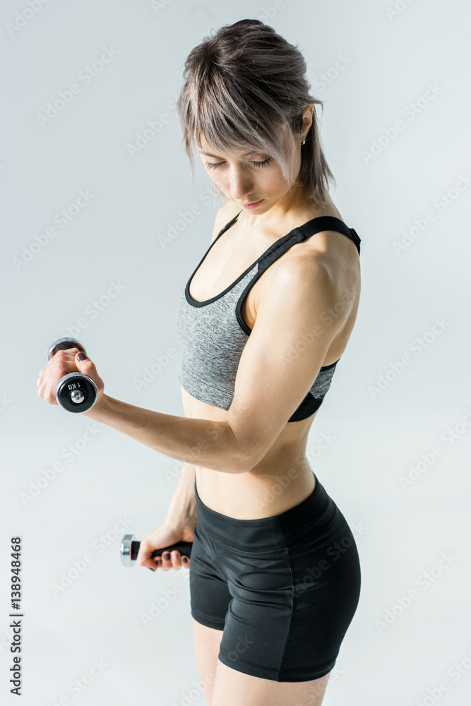 Young sportswoman training with dumbbells and looking at biceps on grey Young sportswoman training with dumbbells and looking at biceps on grey