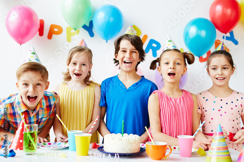 Portrait of five joyous children posing at birthday table with cake on it, smiling and showing their teeth with mouth open