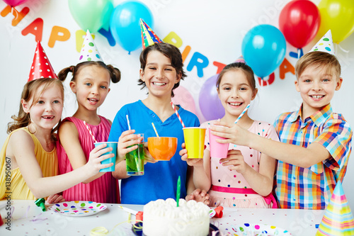 Colorful portrait of five happy kids standing together posing, holding up their drinks in celebration of birthday, looking at camera and smiling