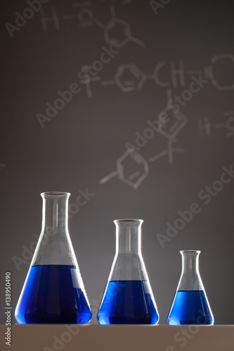 Dark Lab Glassware with chemical formulas in the background, and some sort of fluid.