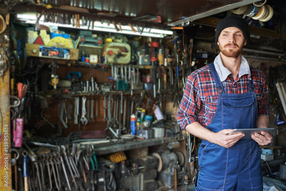 Portrait of handsome bearded mechanic wearing uniform overalls standing holding digital tablet against background of shelves filled with tools in workshop Portrait of handsome bearded mechanic wearing uniform overalls standing holding digital tablet against background of shelves filled with tools in workshop