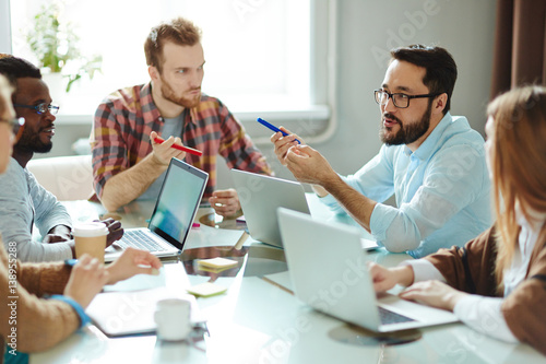 Multi-ethnic team of ambitious designers in casualwear suggesting ideas concerning their new project while sitting together in boardroom
