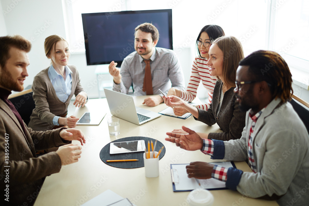 Multi-ethnic group of workers sitting at table in meeting room and sharing ideas concerning new project with their colleagues Multi-ethnic group of workers sitting at table in meeting room and sharing ideas concerning new project with their colleagues
