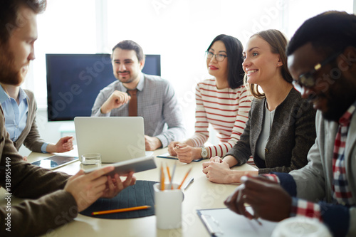 Waist-up portrait of enthusiastic bearded worker holding digital tablet in hands and presenting his project to colleagues, they listening to him with interest