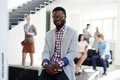 Portrait of smiling African American employee in eyeglasses sitting on back of armchair in office lobby and looking at camera, his colleagues chatting with each other while having break