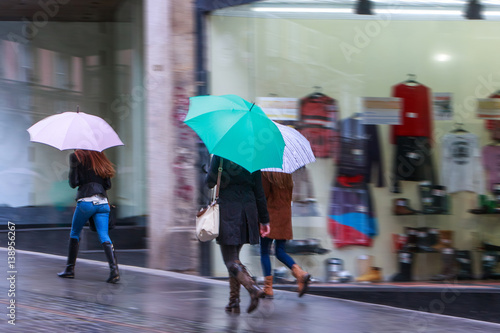 Storm weather. People with umbrellas, pedestrians on the sidewalk in the rain. The storefronts on the city street.