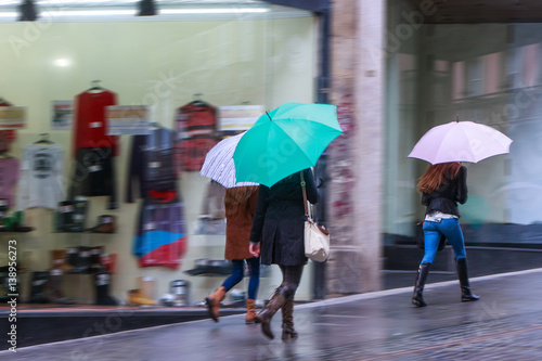 Storm weather. People with umbrellas, pedestrians on the sidewalk in the rain. The storefronts on the city street.