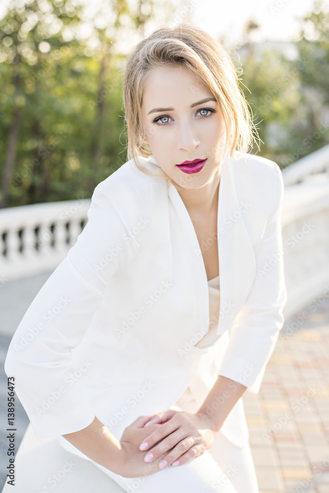 Beautiful blonde girl with red lips in a business white trouser suit, with hair styling and professional makeup on a sunset background. The concept of successful and strong woman close up portrait