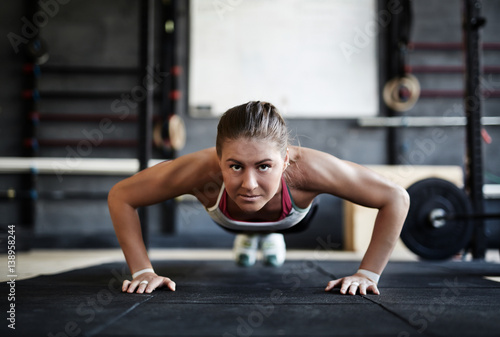 Intense crossfit workout in dark gym: front view of strained young sportswoman performing push us from floor looking at camera