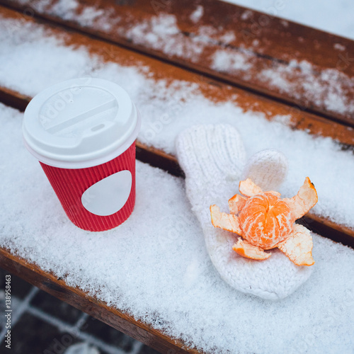 Open madarin lies on white glove behind red paper cup with coffee on snowed bench photo