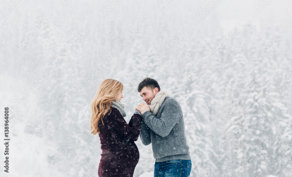 Man holds woman's hands before his mouth while they stand befoe snowy forest Man holds woman's hands before his mouth while they stand befoe snowy forest