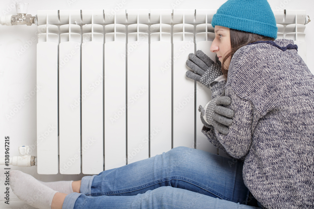 Young beautiful girl froze and warms hands near a radiator, dressed in gloves, a sweater and a hat Young beautiful girl froze and warms hands near a radiator, dressed in gloves, a sweater and a hat