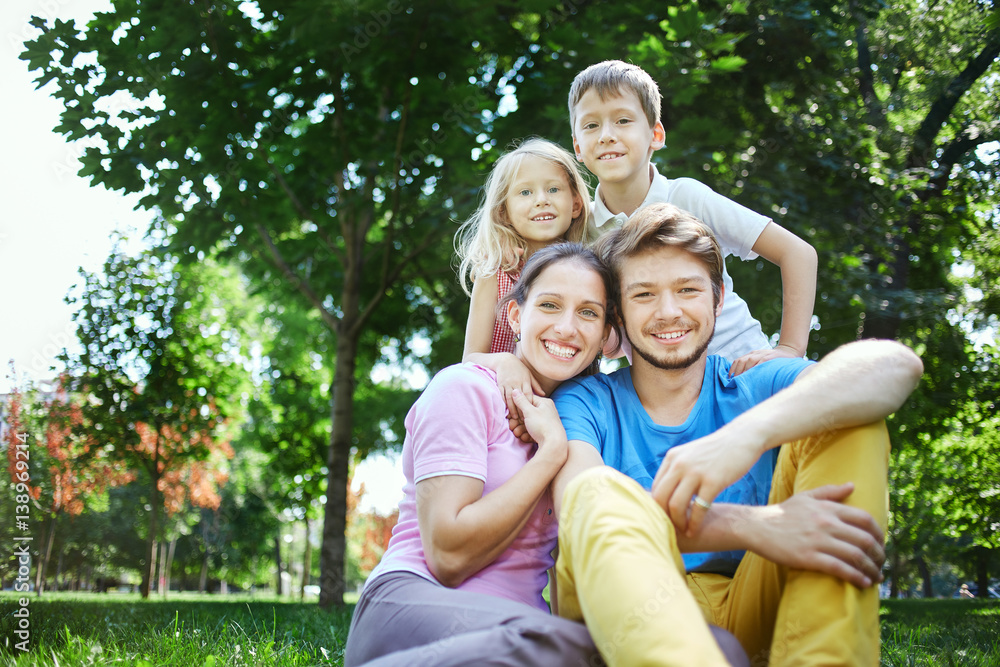 young happy family at noon in the park on the grass. Two young parents and children, boy and girl, lies on the grass and smiling looking at the camera. young happy family at noon in the park on the grass. Two young parents and children, boy and girl, lies on the grass and smiling looking at the camera.