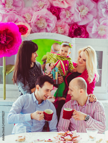 Two men drink tea at dinner table while two ladies play with little girl over them