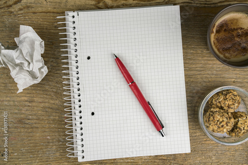 Blank notebook, pen and crumpled paper alongside a mug of hot coffee and cookies on an old wooden desk, overhead close up view