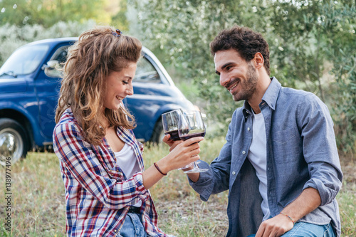 Couple of young lovers drinking red wine from glass goblet at picnic in countryside, Tuscany, Italy. Sitting on lawn among the olive trees on a late summer day, behind them  old blue car © loreanto