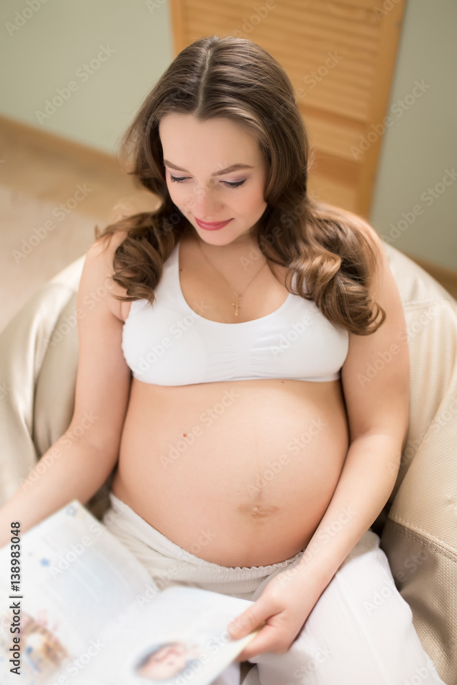Pregnant woman sitting on pouf in home and reading a magazine Pregnant woman sitting on pouf in home and reading a magazine